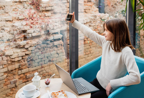 Fille à une table dans un café avec un smartphone et un ordinateur portable prend un selfie. - Photo, image