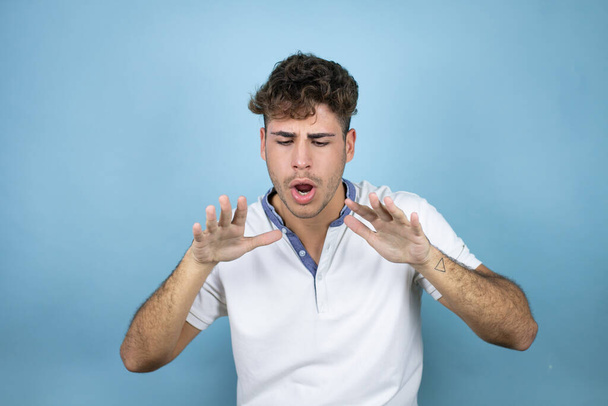 Young handsome man wearing a white t-shirt over blue background shouting and screaming loud down with hands on mouth - Photo, Image