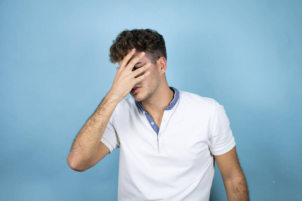 Jonge knappe man draagt een wit t-shirt over blauwe achtergrond gluurend in shock over gezicht en ogen met de hand, kijkend door vingers met beschaamde uitdrukking - Foto, afbeelding