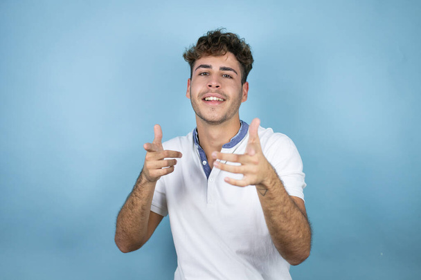 Mladý pohledný muž v bílém tričku na modrém pozadí ukazuje prstem na vás a kameru, usmívá se pozitivně a vesele - Fotografie, Obrázek