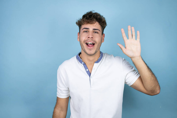 Молодой красивый мужчина в белой футболке на синем фоне показывает и указывает пальцами номер пять, улыбаясь уверенно и счастливо - Фото, изображение