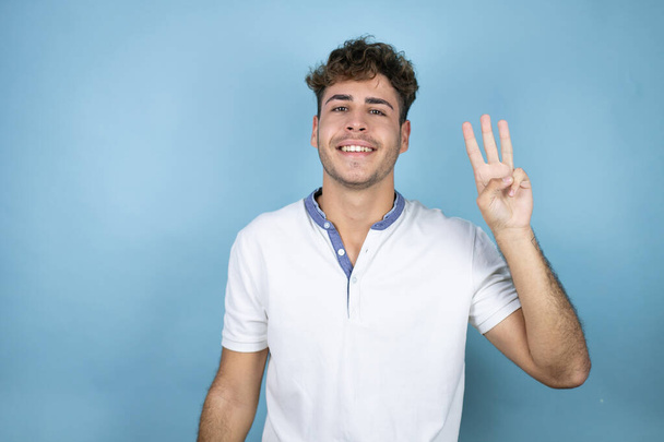 Young handsome man wearing a white t-shirt over blue background showing and pointing up with fingers number three while smiling confident and happy - Foto, immagini