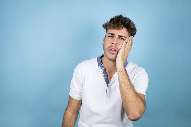 Mladý pohledný muž v bílém tričku přes modré pozadí dotýkání úst s rukou s bolestivým výrazem, protože bolest zubů nebo zubní nemoc na zubech - Fotografie, Obrázek