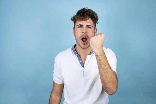 Jonge knappe man draagt een wit t-shirt over een blauwe achtergrond boos en gek razende vuist gefrustreerd en woedend terwijl hij schreeuwt van woede. Woede en agressief concept. - Foto, afbeelding