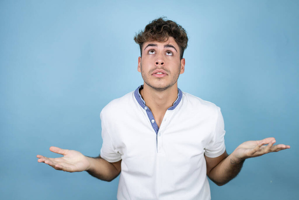 Young handsome man wearing a white t-shirt over blue background clueless and confused expression with arms and hands raised - Photo, Image
