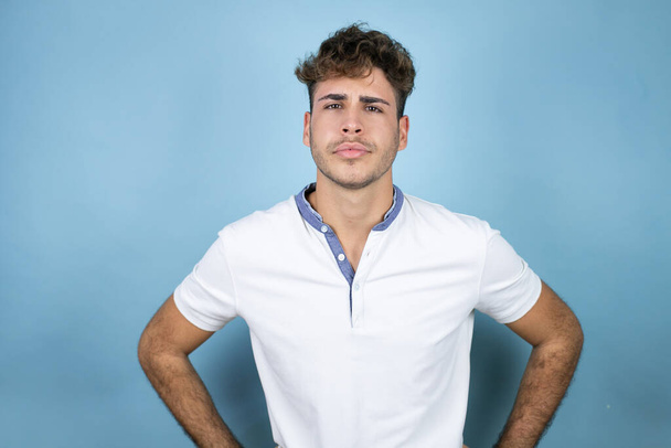 Νεαρός όμορφος άντρας με λευκό μπλουζάκι πάνω από μπλε φόντο σκεπτικός και νευρικός, αποδοκιμάζει την έκφραση στο πρόσωπο με τα χέρια στη μέση - Φωτογραφία, εικόνα