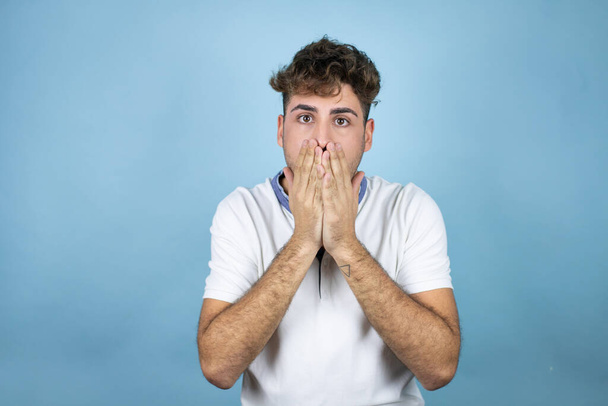 Young handsome man wearing a white t-shirt over blue background with his hands over his mouth and surprised - Foto, afbeelding