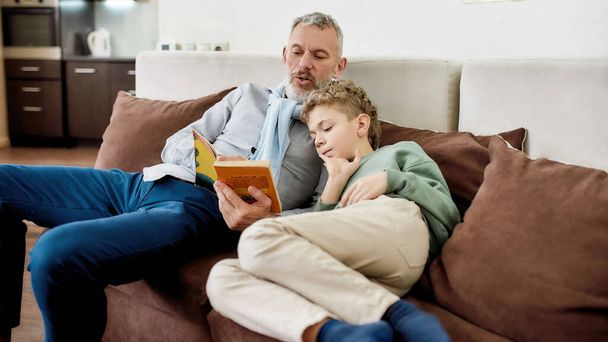 Lets read a story. Grandfather reading a book with his little grandson while relaxing on sofa at home - Foto, Imagem
