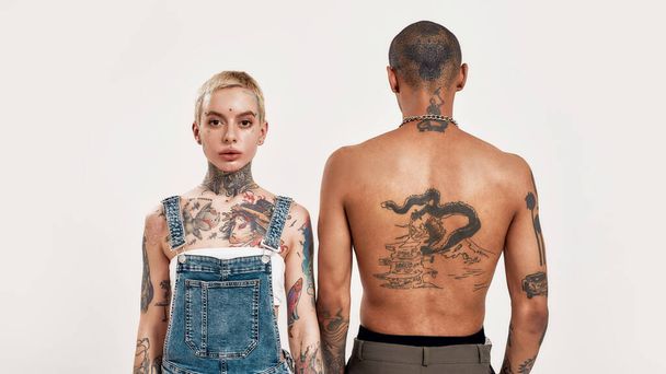 Multicultural relationship. A white pierced woman standing with her face to a camera next to a dark-skinned topless male with tattoos standing with his back to a camera - Foto, Imagem