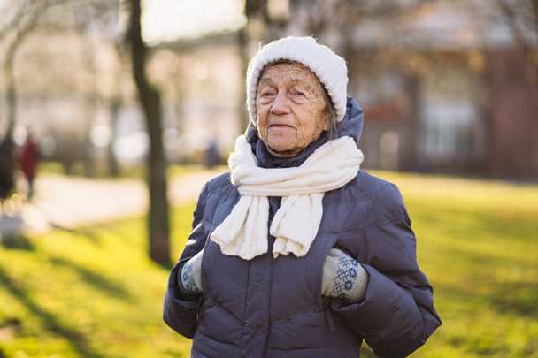 Portrait Caucasian senior woman with gray hair and deep wrinkles 90 years old posing in warm clothes, white knitted scarf and hat in park, sunny frosty weather. Active old age, walking elderly female. - Photo, Image