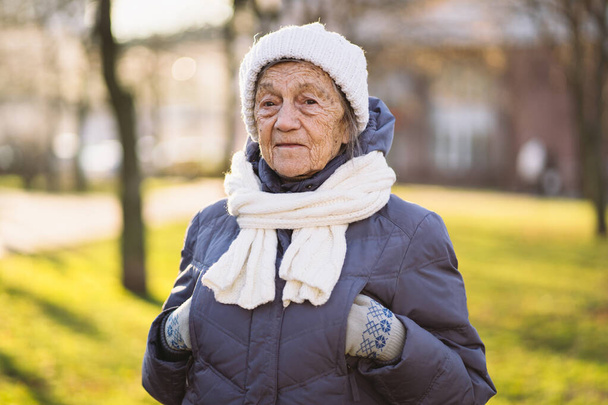 Joyful mature lady expressing positive emotions in winter knitted white hat and scarf in blue coat in city park. Senior woman cold season outside. Excellent health in retirement. retired 90 years old. - Photo, Image