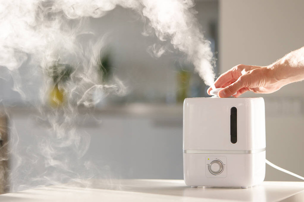 Hand turn on aroma oil diffuser on the table at home, steam from the air humidifier, free space. Ultrasonic technology, increase in air humidity indoors, comfortable living conditions, moisture.  - Photo, Image