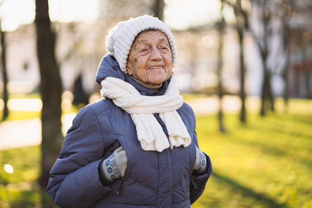Retrato mujer mayor caucásica con el pelo gris y las arrugas profundas 90 años posando en ropa de abrigo, bufanda de punto blanco y sombrero en el parque, tiempo helado soleado. La vejez activa, caminar anciana mujer. - Foto, Imagen