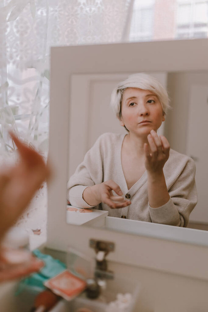 Young millennial woman is dressing up. Wearing earrings. Beauty routine. Make up artist. Mascara, lipstick, blushes. Pastel colors. Dressing vanity mirror. Pretty blonde lady. White bedroom light. - Foto, Bild