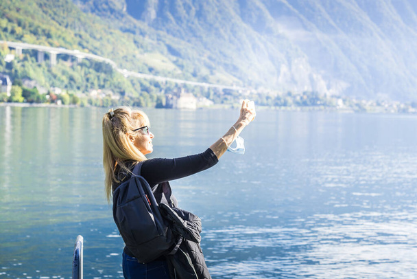 Mujer rubia con gafas de sol y una bolsa haciendo una selfie en un lago rodeado de una ciudad y colinas con una luz dura que viene del sol. Montreux en el lago Geneve - Foto, Imagen