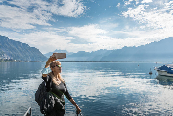 Donna bionda con gli occhiali da sole una maschera nel braccio un selfie in un lago con una barca circondata da una città e colline con una luce dura proveniente dal sole. Montreux nel lago di Geneve - Foto, immagini