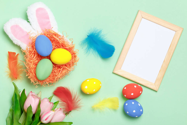 Easter eggs in a nest, bunny ears, flowers and a frame with copy space on a mint green background. - Zdjęcie, obraz