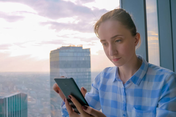 Retrato de la mujer usando el dispositivo de teléfono inteligente contra la vista del paisaje urbano a través de la ventana - Foto, Imagen