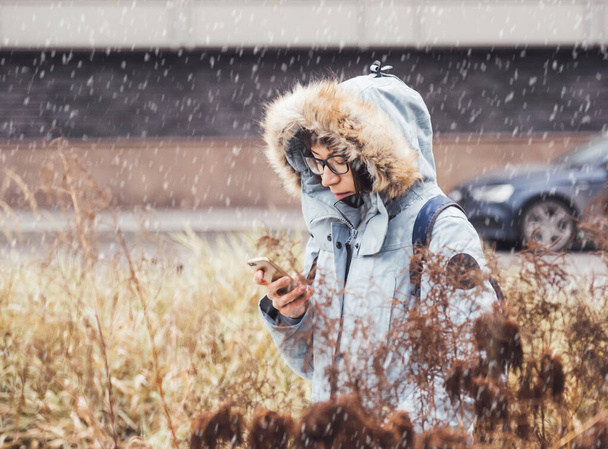 Smiling woman in light blue warm jacket is texting someone on smartphone. Online communication via LTE or Wi-fi. Wireless cellular technology. Autumn season in town. Snowfall. - Foto, Imagem