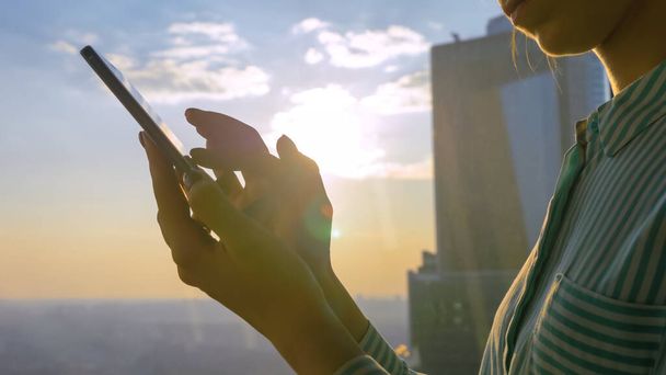 Woman using smartphone against sunset cityscape through window of skyscraper - Photo, Image