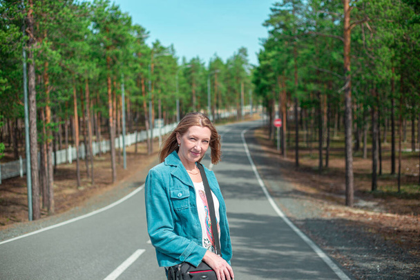 blonde woman with long hair stands on the road in the forest, traveling on a summer sunny day - Photo, Image