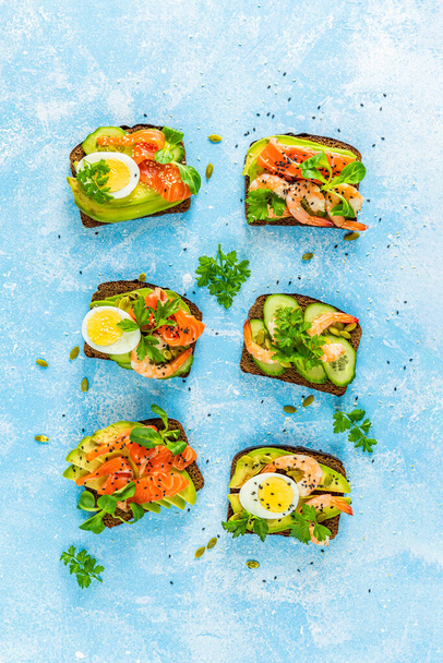 Healthy snack or appetizer toast with salmon, avocado, shrimps, eggs, cucumber, lambs lettuce or corn salad and parsley. Top view - Photo, Image