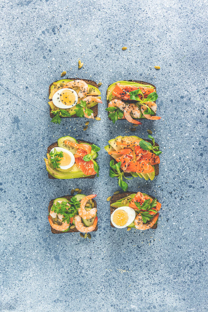 Healthy snack or appetizer toast with salmon, avocado, shrimps, eggs, cucumber, lambs lettuce or corn salad and parsley. Light grey table surface - Photo, Image