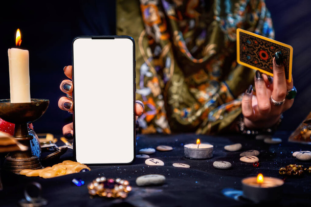 De waarzegster heeft een Tarotkaart en een smartphone met een wit scherm. Maak je niet druk. Een close-up. Het concept van waarzeggerij, magie en esoterie. - Foto, afbeelding
