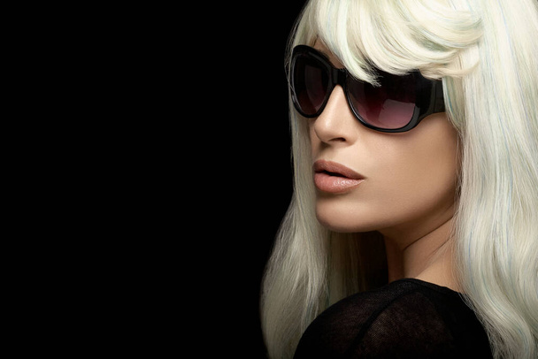 portrait de mode d'une jolie femme blonde aux longs cheveux blancs argentés, portant des lunettes de soleil noires, regardant en arrière sur l'épaule. gros plan isolé sur fond noir avec espace de copie pour le texte - Photo, image