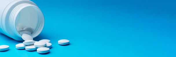 Witte pillen zijn verspreid in de buurt van de ondersteboven pot op een blauwe achtergrond. Calcium vitaminen zijn verspreid. Botpillen in de buurt van de witte container. - Foto, afbeelding