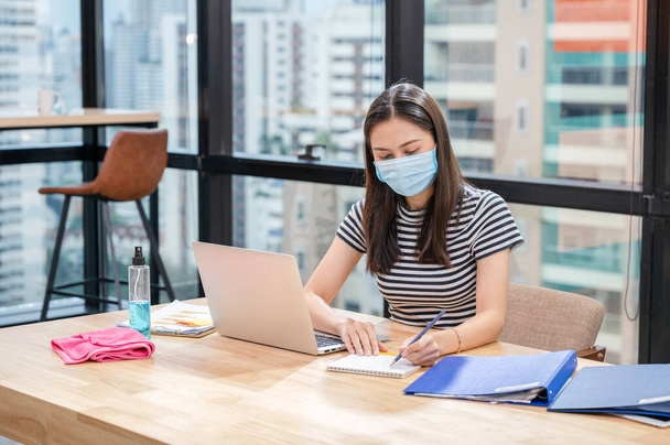 Young asian woman in casual with face mask and note of schedule on the calendar. New normal office with desk, laptop, documents and alcohol spray while pandemic of coronavirus, covid-19 - Photo, Image