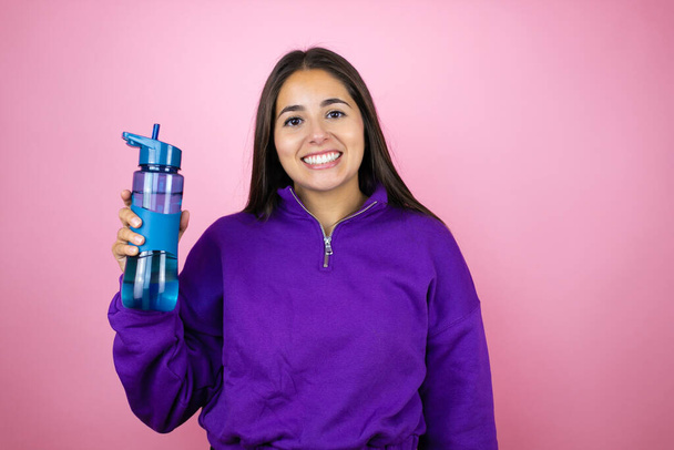 Young beautiful woman wearing sweatshirt holding a water bottle over isolated pink background with a happy face standing and smiling with a confident smile showing teeth - Фото, изображение