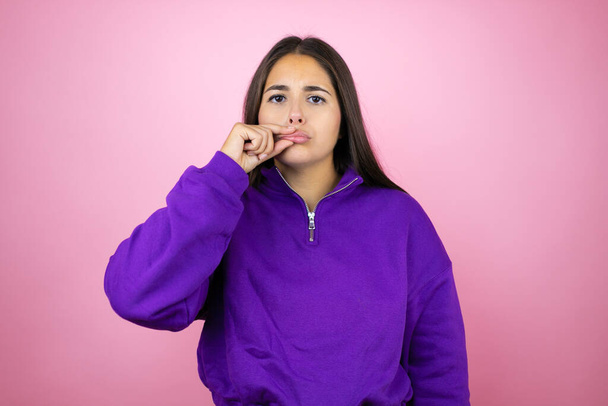Jeune belle femme portant un sweat-shirt sur fond rose isolé bouche et lèvres fermées comme zip avec les doigts. Secret et silencieux, tabou parlant - Photo, image