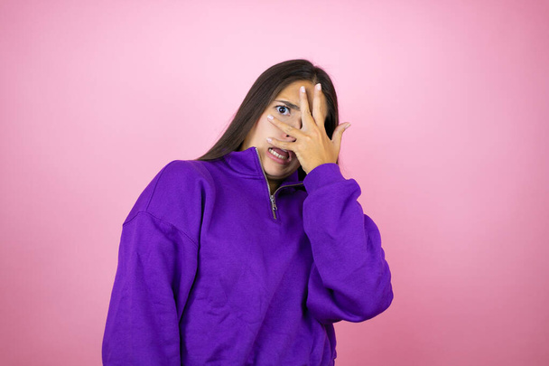 Jonge mooie vrouw dragen sweatshirt over geïsoleerde roze achtergrond gluren in shock bedekking gezicht en ogen met de hand, kijken door vingers met beschaamde uitdrukking - Foto, afbeelding