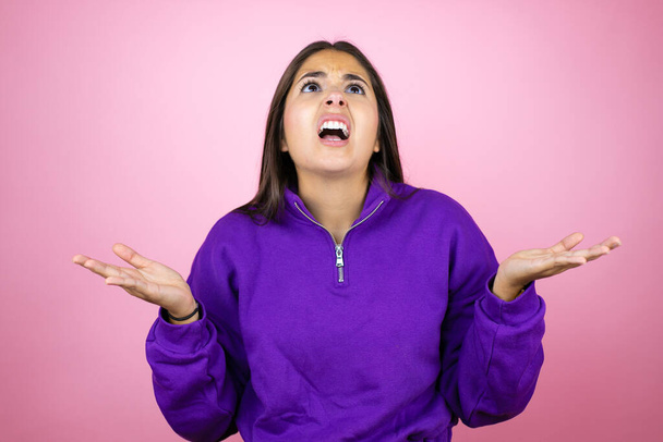 Young beautiful woman wearing sweatshirt over isolated pink background clueless and confused expression with arms and hands raised - Photo, Image