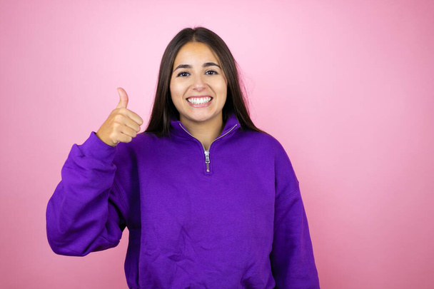 Young beautiful woman wearing sweatshirt over isolated pink background success sign doing positive gesture with hand, thumb up smiling and happy. cheerful expression and winner gesture. - Photo, Image