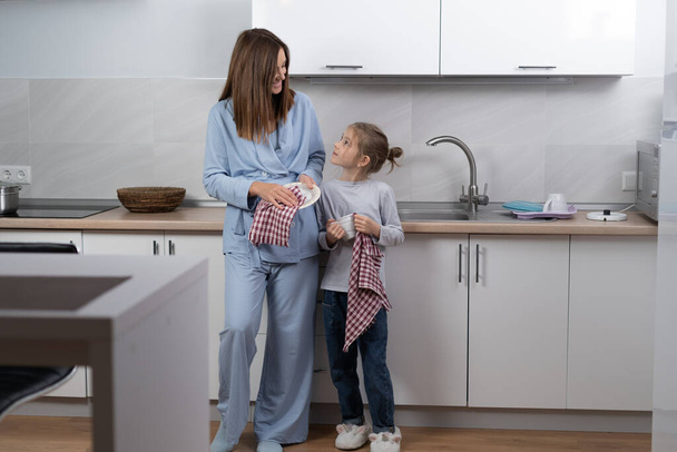 Όμορφη νεαρή γυναίκα με την κόρη της στην κουζίνα σκουπίζει τα πιάτα, κοιτάζει ο ένας τον άλλο και χαμογελά ενώ καθαρίζει την κουζίνα - Φωτογραφία, εικόνα