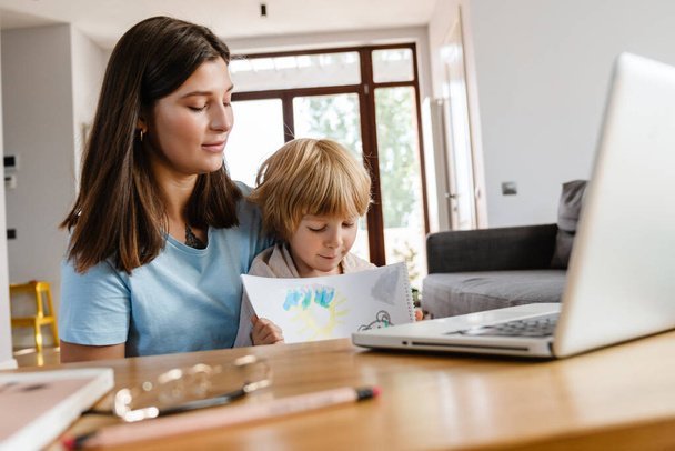 Image of beautiful woman at home with her son showing his drawings by videocall by laptop computer - Photo, image