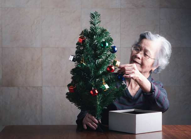 Portré egy idős ázsiai nőről, aki karácsonyfát díszít, miközben otthon ül egy nappaliban. Helyet a szövegnek. Fogalom az idősek és a karácsony és boldog új évet fesztivál. - Fotó, kép