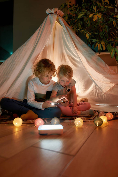 Two little kids, brother and sister using tablet pc while sitting on a blanket in a teepee made with bedsheets at home - Foto, Imagen