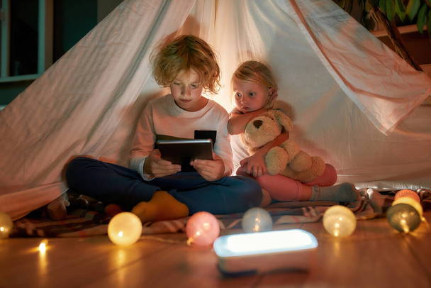 Adorable boy using tablet pc while spending time with his little sister, sitting on a blanket in a hut made with bedsheets at home - Foto, Imagen