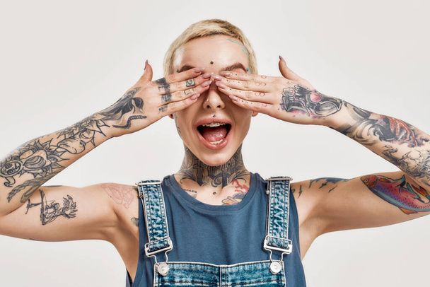 Emotions and postures. A white pierced and tattooed woman covering her eyes with her palms wearing a denim overall with her mouth widely open and smiling - Zdjęcie, obraz