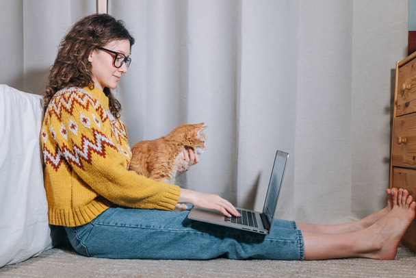 Woman works at home on a laptop sitting on carpet with her red cat, remote work from home, freelance. - Foto, Imagen