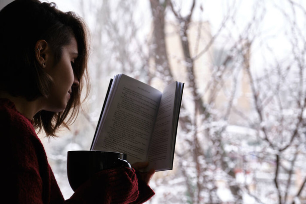 Eine junge Frau hält eine heiße Kaffee- oder Teetasse in der Hand und liest an einem verschneiten Wintertag am Fenster ein Buch. Gemütliches Zuhause-Konzept. - Foto, Bild