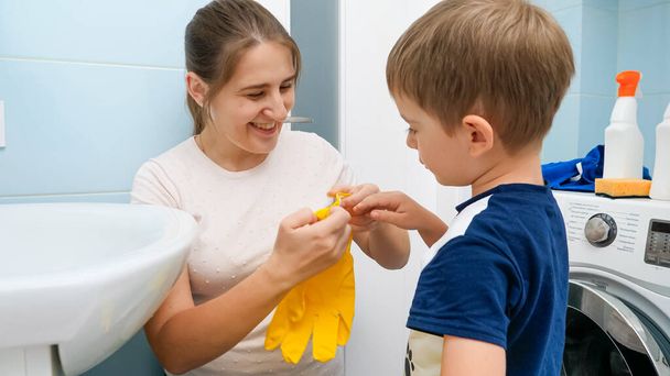 Lächelnde junge Mutter, die ihrem kleinen Sohn hilft, gelbe Gummihandschuhe anzuziehen, bevor sie im Badezimmer Hausarbeit erledigt. Kinder helfen Eltern bei Hausarbeit und Alltag. - Foto, Bild