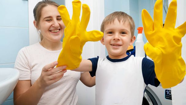 Vtipný rozesmátý chlapec ve žlutých gumových rukavicích, smějící se a šklebící se, než udělá domácnost a uklízí koupelnu se svou matkou. Rodinné foing domácí práce a baví - Fotografie, Obrázek