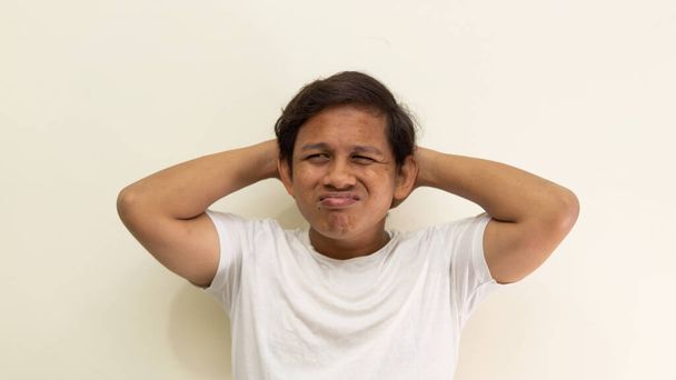 Verward en verbijsterd gezicht uitdrukking met de hand achter hoofd van jonge Aziatische Maleisische man met witte t-shirt op geïsoleerde witte achtergrond. Verwarrend handgebaar. - Foto, afbeelding