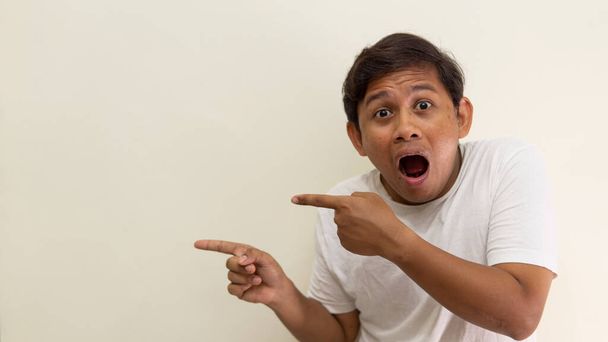 Wow en geschokt gezicht uitdrukking van jonge Aziatische Maleisische man in wit t-shirt met de hand wijzend op lege ruimte op geïsoleerde witte achtergrond. - Foto, afbeelding