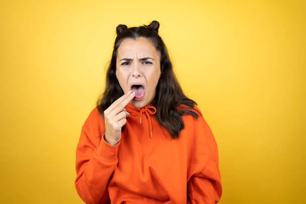 Young beautiful woman wearing sweatshirt over isolated yellow background disgusted with her hand inside her mouth - 写真・画像