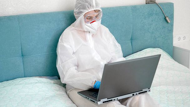 ベッドに座ってノートパソコンで作業する医療用スーツとマスクマスクマスクを身に着けているフリーランスの労働者は、ロックダウンとcovid-19パンデミックの間に - 写真・画像